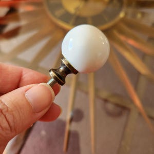 May include: A white, spherical cabinet knob with a bronze-colored base and screw. The knob is held in a person's hand, with a blurred background featuring a gold-colored clock.