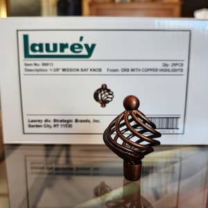 May include: A bronze Mission Bay knob with copper highlights. The knob has a decorative cage-like design and a round top. The box behind the knob reads "laurey" and "1-3/8" MISSION BAY KNOB".