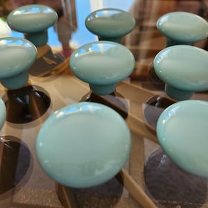 May include: A collection of ten round, smooth, light blue cabinet knobs arranged in a grid pattern. Each knob features a glossy, reflective surface with a simple, circular top and a cylindrical base, providing a uniform appearance for furniture.