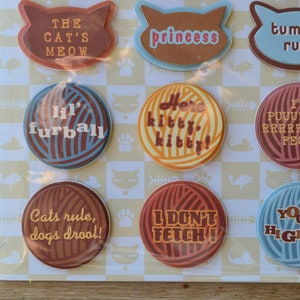 May include: Nine round, colorful buttons with cat-themed sayings. The buttons feature various colors and patterns, including brown, blue, yellow, and red. The sayings on the buttons include "The Cat's Meow", "Princess", "Here Kitty Kitty", "Lil' Furball", "Tummy Rub", "I'm Purrrrrrrrfect", "Cats Rule, Dogs Drool", "I Don't Fetch", and "Your Highness".