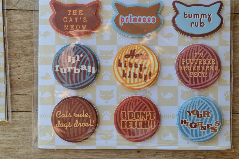 May include: Nine round, colorful buttons with cat-themed sayings. The buttons feature various colors and patterns, including brown, blue, yellow, and red. The sayings on the buttons include "The Cat's Meow", "Princess", "Here Kitty Kitty", "Lil' Furball", "Tummy Rub", "I'm Purrrrrrrrfect", "Cats Rule, Dogs Drool", "I Don't Fetch", and "Your Highness".