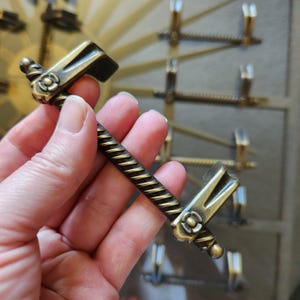 May include: Antique brass-toned cabinet pull with a decorative floral design. The pull has a twisted handle and a rectangular grip. The hardware is likely for a drawer or cabinet door.