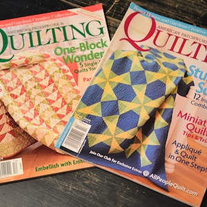 May include: Two copies of the American Patchwork & Quilting magazine. The cover of the magazine on the left features a quilt with a red, pink, and green color scheme. The cover of the magazine on the right features a quilt with a blue, yellow, and white color scheme. The text on the cover of the magazine on the left reads "Easy Gifts to Make" and "One-Block Wonder". The text on the cover of the magazine on the right reads "Stunning Solids" and "Miniature Quilts".