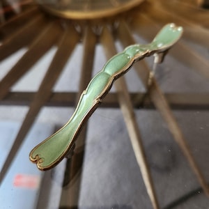 May include: A decorative cabinet pull handle with a light green enamel finish and gold-colored trim. The handle has a curved, elongated shape with a small foot. The handle is likely made of metal.