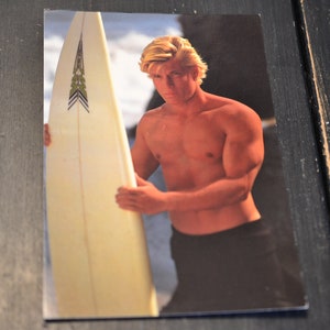 May include: A young man with blond hair is holding a white surfboard with a green and black design. He is wearing black swim trunks and is standing in the ocean.