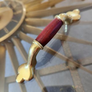 May include: A close-up of a cabinet handle. It features a dark red wooden grip and gold-coloured metal accents. The handle has a curved design with a decorative end. The background shows a portion of a gold-coloured clock.