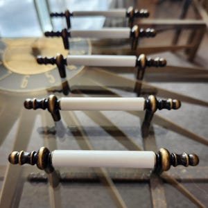 May include: Five white ceramic drawer pulls with gold metal accents. The pulls are arranged in a row, with the ends of the pulls pointing towards the center of the image.