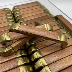 May include: A collection of rectangular wooden drawer pulls featuring a smooth, medium-brown wood grain finish and antique brass-toned metal end caps. Two of the pulls are positioned in a crossed arrangement over the others, which are aligned.
