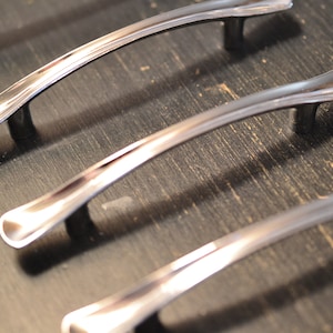 May include: Four silver cabinet pulls with a curved design. The pulls are arranged in a row on a dark wood surface.