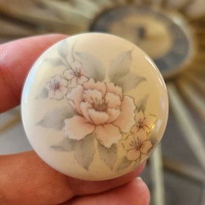 May include: A round, cream-colored ceramic trinket box with a floral design. The flowers are pink, white, and gray.