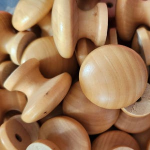 May include: A close-up of a pile of unfinished wooden knobs and pulls. The knobs are circular, and the pulls have a T-shape. The wood is a light, natural colour with visible grain. These are likely for furniture or cabinet making.