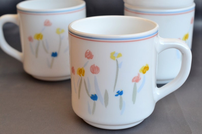May include: Three white ceramic mugs with a floral design. The mugs have a blue, pink, yellow, and green floral pattern. The mugs have a blue and red rim.
