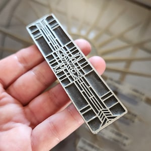 May include: A silver metal door handle with a geometric design. The handle is rectangular in shape and features a raised pattern of lines and squares.