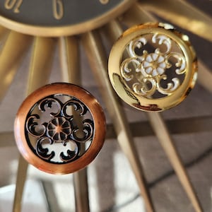 May include: Two decorative clock accents. One is copper-toned with a black filigree design, and the other is gold-toned with a white and gold filigree design. The clock accents are circular and ornate.