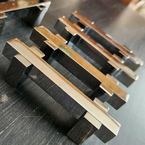 May include: Close-up of several silver and black cabinet pulls. The pulls have a rectangular design with a silver bar across the top and black supports. The pulls are arranged on a dark surface.