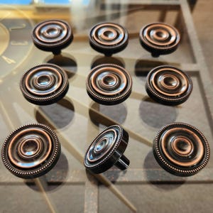 Peut inclure: Neuf boutons de meuble de couleur bronze avec un design circulaire. Chaque bouton présente des cercles concentriques et un bord perlé. Les boutons sont disposés sur une surface réfléchissante, mettant en valeur leur finition métallique et leurs détails complexes.
