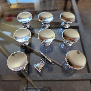 May include: Ten silver-toned cabinet knobs with a round, slightly domed top and a short, cylindrical base. One knob is shown with the top removed, revealing the base.