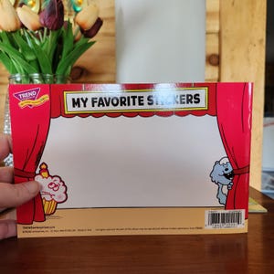 May include: A red and white sticker album with the text "MY FAVORITE STICKERS". The album features cartoon illustrations of a cupcake and a blue creature. The album is held in front of a wooden surface.