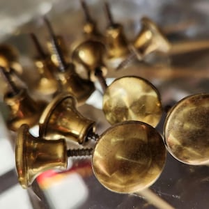 Peut inclure: Gros plan de plusieurs boutons de meubles anciens en laiton avec vis. Les boutons ont une variété de formes, y compris des designs arrondis et en forme de cloche. Le laiton a une teinte dorée chaude, et les vis sont visibles.
