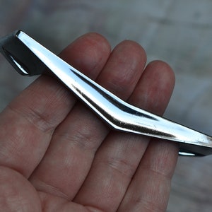 May include: A shiny, chrome-finished cabinet pull handle. The handle has a sleek, modern design with a curved shape and a polished surface. It is held in a hand, showcasing its size and form.