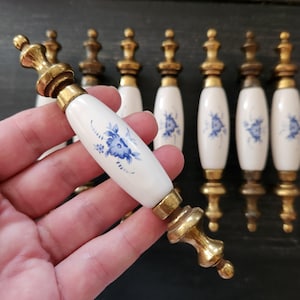 Peut inclure: Un ensemble de sept poignées de tiroir avec un centre en céramique blanche orné d'un motif floral bleu et des extrémités en métal doré. Chaque poignée possède un corps cylindrique en céramique et des embouts décoratifs en métal.