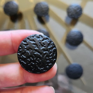 Peut inclure: Gros plan sur un bouton rond noir texturé, tenu entre les doigts. Le bouton présente un motif en relief organique. Plusieurs autres boutons similaires sont flous en arrière-plan, suggérant un ensemble de quincaillerie décorative.