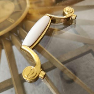 May include: A gold-toned cabinet pull with a white, oval-shaped center. The pull has a curved design with decorative scrollwork at each end. The hardware is likely for a drawer or cabinet door.