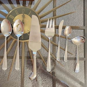 May include: A set of seven silver-toned utensils, including a serving knife, a serving spoon, a butter knife, a fork, and three teaspoons. The utensils are arranged on a gold-colored surface with a clock face in the background.