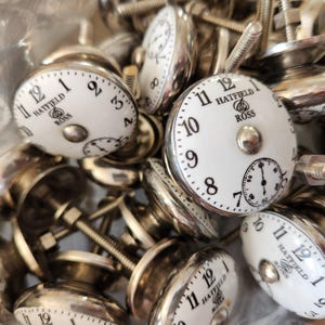 May include: A collection of white and silver cabinet knobs with clock faces. The knobs feature black numerals and the text "HATFIELD ROSS". The knobs have a vintage, antique aesthetic and are ideal for furniture restoration projects.