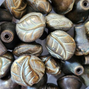 May include: A selection of bronze-coloured cabinet knobs. The knobs have a leaf design and a vintage appearance. Some are round, others cylindrical. The knobs are likely made of metal and would suit a traditional or antique-style interior.