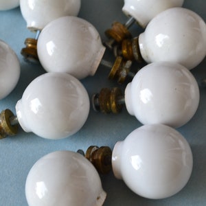 May include: A collection of white ceramic cabinet knobs with brass hardware. The knobs are round with a glossy finish and are scattered on a light blue surface. The hardware includes screws and washers.