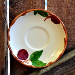 May include: A white ceramic saucer with a brown rim and a brown, green, and red floral design. The saucer has a small depression in the center for a cup.