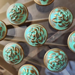 May include: A collection of round cabinet knobs featuring a raised floral design. Each knob has a bright turquoise finish with distressed bronze-colored highlights on the petals and edges, creating a textured appearance across the circular surface.