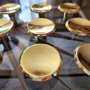 May include: A collection of circular cabinet knobs with a polished gold finish. Each knob features a smooth, slightly concave top surface and a reflective metallic sheen, arranged in a grid pattern to showcase the uniform design and reflective quality.