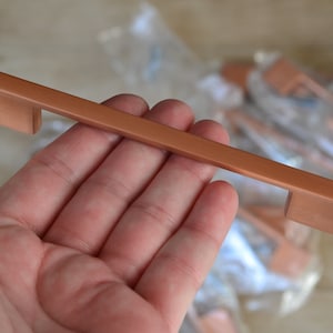 May include: A copper cabinet pull with a rectangular shape and a cut-out handle. The pull is being held in a hand.