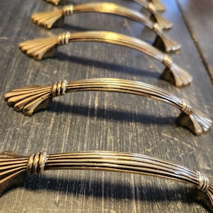 May include: A set of five vintage brass cabinet handles. Each handle has a curved, ribbed design with decorative fan-shaped ends and a central, textured grip. The handles are arranged on a dark, textured surface.