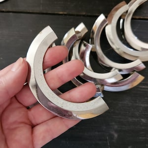 May include: A collection of silver-toned, crescent-shaped metal jewelry components. The pieces have a curved design with a polished finish, and are held in a hand against a dark background. The components appear to be for jewelry making.