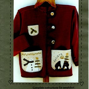 May include: A burgundy sweatshirt with a star-shaped collar and appliqued pockets. The pockets feature snowman and Santa Claus designs, with the word "believe" on one. The text "Heart to Hand" is also visible.