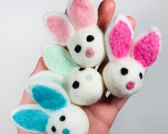 May include: Four small needle-felted white bunny heads with fuzzy textures. Each bunny features two upright ears in distinct colors: one pair in soft pink, one in bright pink, one in mint green, and one in teal. Each has two black bead eyes and a nose.