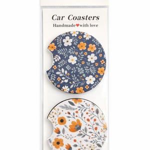 May include: Two floral car coasters. One coaster has a navy blue background with white and orange flowers. The other coaster has a white background with orange flowers and gray leaves. The packaging reads "Car Coasters Handmade with love."