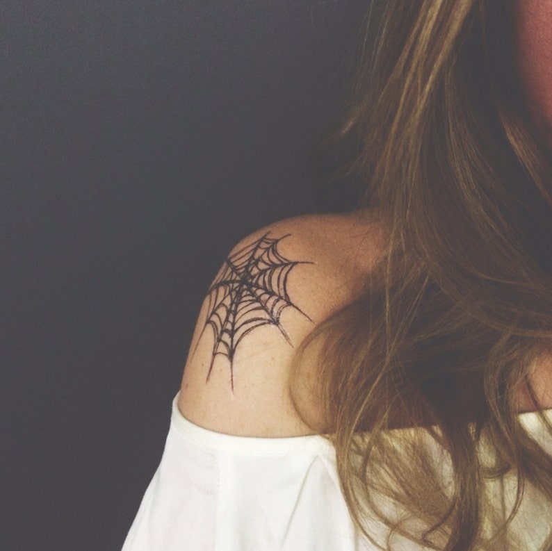 May include: A black ink spider web tattoo design positioned on the shoulder. The tattoo features fine, dark lines forming a classic web pattern with a central point and radiating strands, displayed on skin above a white off-the-shoulder garment.