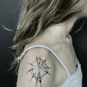 May include: A black ink spider web tattoo design positioned on the shoulder area. The tattoo features fine, dark lines forming a classic web pattern with a central point, displayed on skin with a light grey tank top strap visible over the shoulder.