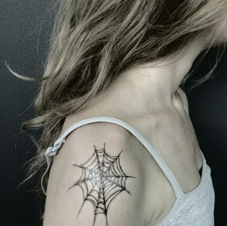 May include: A black ink spider web tattoo design positioned on the shoulder area. The tattoo features fine, dark lines forming a classic web pattern with a central point, displayed on skin with a light grey tank top strap visible over the shoulder.