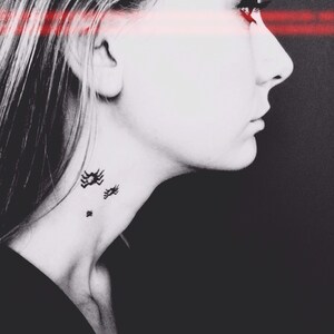 May include: A black and white profile portrait of a person with three small black spider tattoos on the side of the neck. A horizontal red light beam crosses the eyes, creating a high-contrast effect against the dark background.