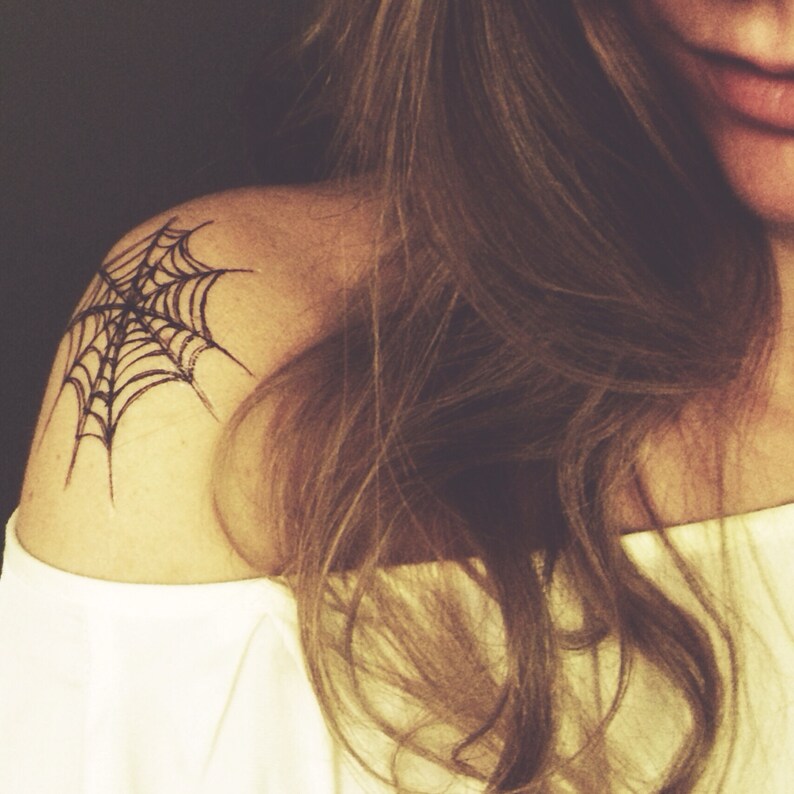 May include: A black ink temporary tattoo design featuring a detailed spider web pattern applied to the shoulder area. The web consists of radiating lines connected by concentric arcs, creating a geometric structure on the skin surface.