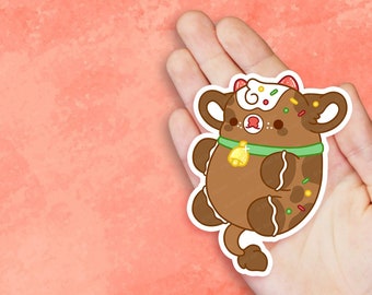 May include: A brown gingerbread cow sticker with white icing details, sprinkles, and a green collar with a gold bell. The sticker is held in a hand against a peach-colored background.