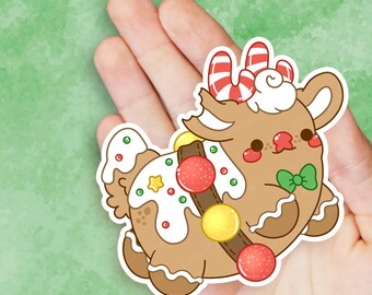 May include: A gingerbread reindeer sticker with white icing, red and green sprinkles, and a candy cane on its antlers. The reindeer is wearing a brown belt with yellow and red candy ornaments.