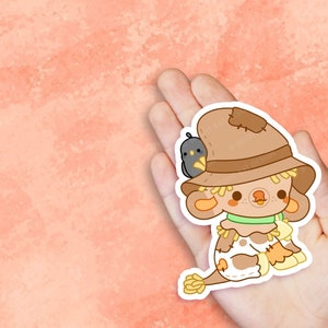 May include: A cute cartoon scarecrow with a brown hat and a small black bird perched on top. The scarecrow has a green collar and a white and brown patterned body. The scarecrow is sitting with its legs crossed and has a friendly expression.