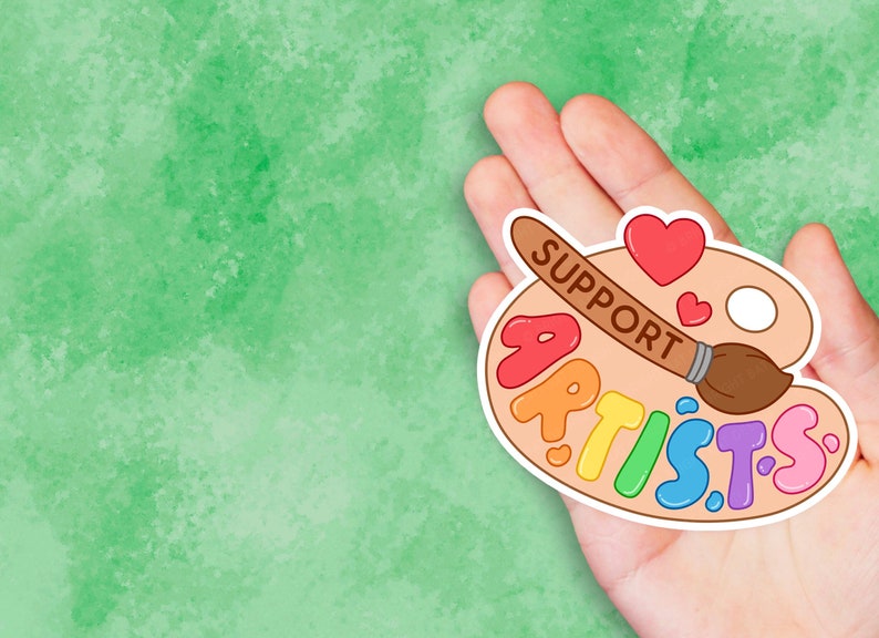 K&ouml;nnte beinhalten: Eine Hand h&auml;lt einen Aufkleber mit einer Farbpalette und den Worten "SUPPORT ARTISTS" in Regenbogenfarben. Die Palette hat einen Pinsel und ein Herz.