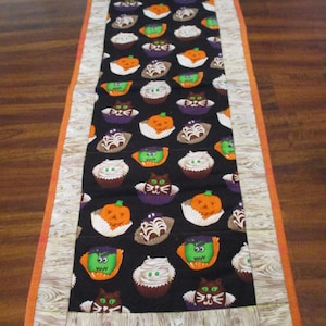 May include: A rectangular Halloween-themed table runner. The center features a black background with repeating cupcake designs, including pumpkins, cats, and ghosts. The border is a light wood grain pattern with an orange trim. The date "09/04/2025" is visible.